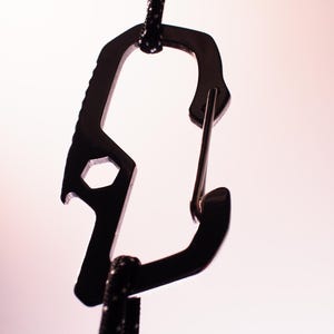 Op de afbeelding: Een zwarte metalen karabijnhaak met een flesopener en een moersleuteltool eraan bevestigd. De karabijnhaak is bevestigd aan een zwart koord.