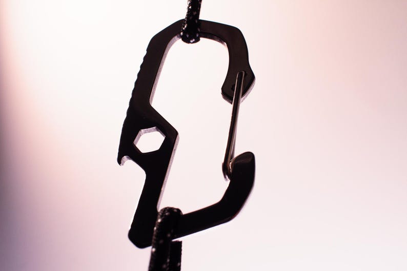 Op de afbeelding: Een zwarte metalen karabijnhaak met een flesopener en een moersleuteltool eraan bevestigd. De karabijnhaak is bevestigd aan een zwart koord.