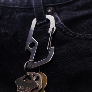 Op de afbeelding: Een zilveren multitool-karabiner met een flesopener en een sleutelhanger. De karabiner is bevestigd aan een sleutelbos.