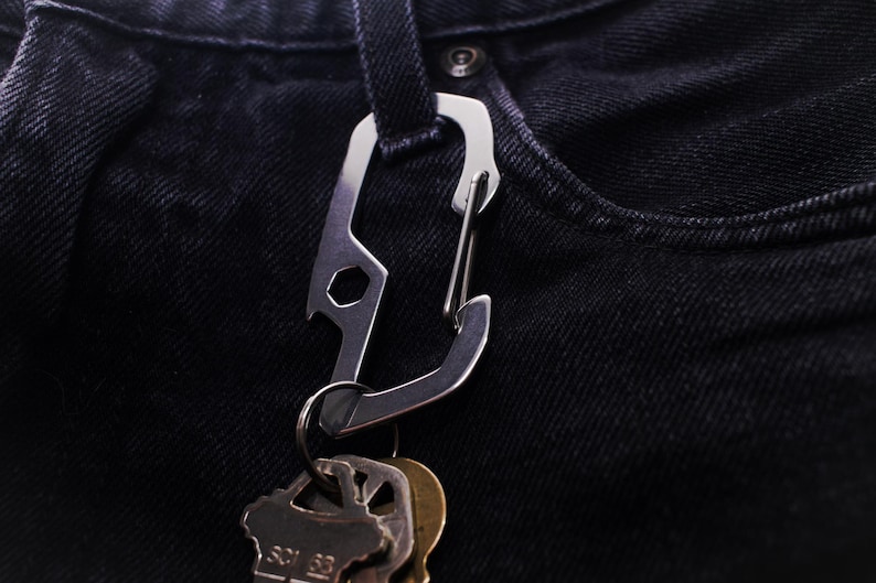 Op de afbeelding: Een zilveren multitool-karabiner met een flesopener en een sleutelhanger. De karabiner is bevestigd aan een sleutelbos.