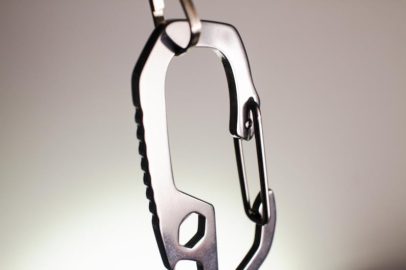Op de afbeelding: Een zilveren metalen karabijnhaak met een ingebouwde flesopener en sleutel. De karabijnhaak heeft een getextureerde grip en een veerbelaste sluiting.