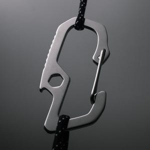 Op de afbeelding: Een zilveren metalen karabijnhaak met een flesopener en een moersleuteltool eraan bevestigd. De karabijnhaak is bevestigd aan een zwart koord.