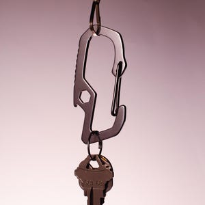 Op de afbeelding: Een zilveren karabijnhaak met een flesopener bevestigd aan een ketting. Een sleutelbos is bevestigd aan de karabijnhaak.