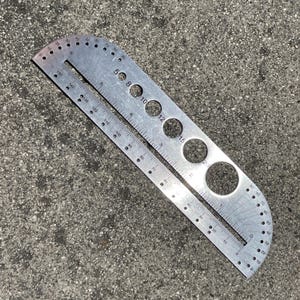 May include: A silver metal protractor with a ruler and multiple circular holes of varying sizes. The tool has degree markings from 0 to 180 and inch measurements. The tool is designed for precise measurements and angles.