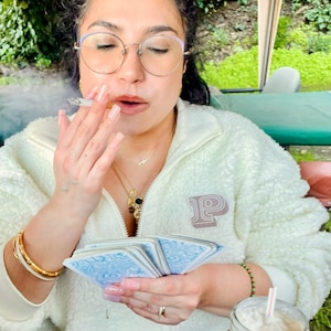 Op de afbeelding: Een persoon draagt een witte fleecejas met een "P"-logo en houdt een pak tarotkaarten met blauwe ontwerpen vast. De persoon rookt een sigaret en draagt gouden sieraden, waaronder een bril en armbanden. Een drankje met een rietje staat op de voorgrond.