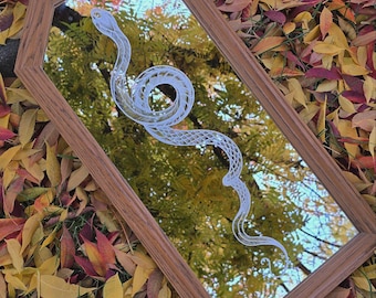 May include: A wooden framed mirror with a white snake etched into the glass. The mirror reflects a tree with fall foliage.