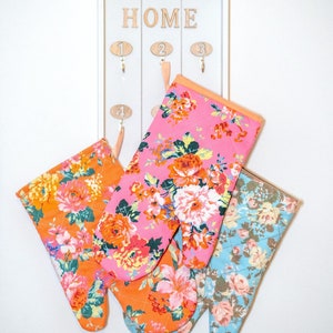 Può includere: Quattro presine floreali nei toni dell'arancione, del rosa e del blu sono appese a un portachiavi bianco a parete. Le presine presentano un design trapuntato con un vivace motivo floreale. Il portachiavi ha la scritta "HOME" in alto.