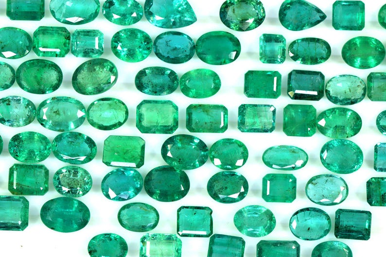 May include: A collection of emerald green gemstones of various shapes and sizes, including oval, rectangular, and square cuts. The gemstones are arranged on a white background.