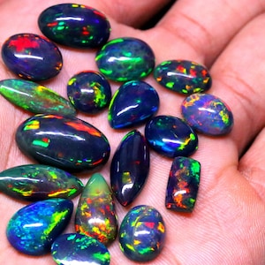 May include: A collection of black opal gemstones with iridescent green, blue, and red flashes. The opals are various shapes and sizes, including oval, teardrop, and rectangular.