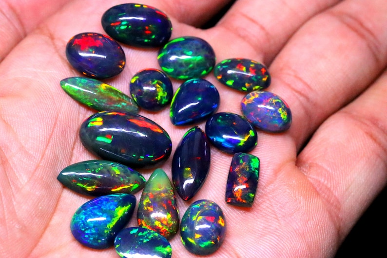 May include: A collection of black opal gemstones with iridescent green, blue, and red flashes. The opals are various shapes and sizes, including oval, teardrop, and rectangular.