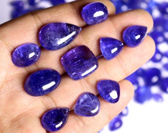 May include: A collection of polished, deep blue gemstones in various shapes, including oval, teardrop, and square. The gems are held in a hand, with a blurred background of similar stones.