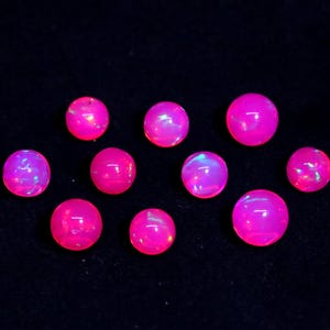 Op de afbeelding: Tien ronde, roze opaal cabochons met iriserende kleurflitsen. De opalen zijn in een losse cluster op een zwarte achtergrond gerangschikt.