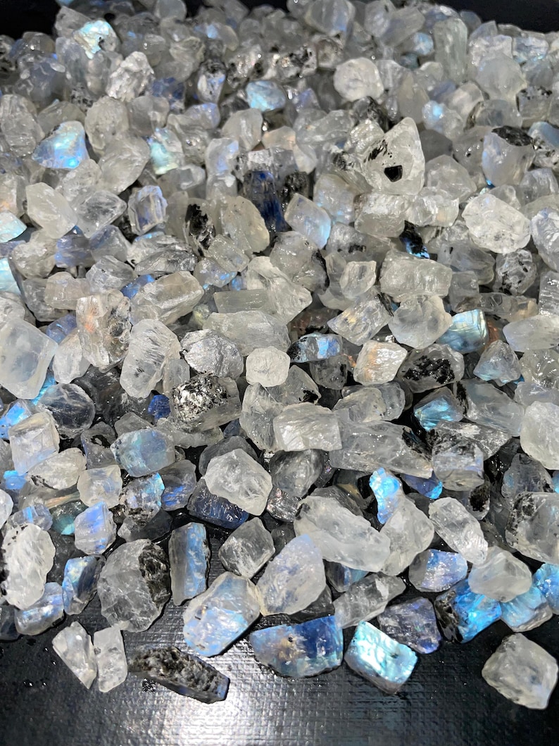 May include: A pile of raw, uncut moonstone crystals. The stones are primarily clear and translucent, with flashes of blue and iridescent color. Some stones have dark inclusions. The crystals vary in size and shape.
