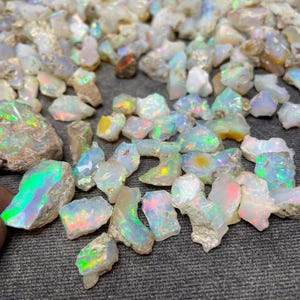 Op de afbeelding: Een verzameling ruwe, witte opaal edelstenen met iriserende regenboogkleuren. De opalen zijn in verschillende maten en vormen, en sommige hebben een ruwe, natuurlijke textuur.