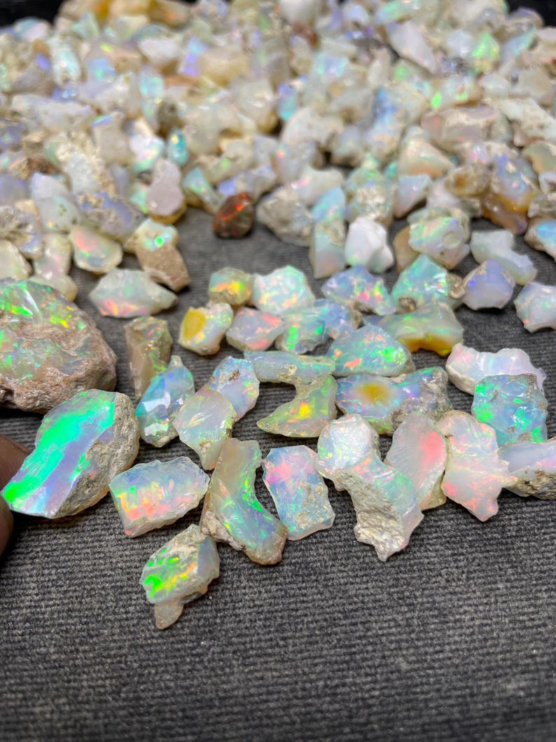 Op de afbeelding: Een verzameling ruwe, witte opaal edelstenen met iriserende regenboogkleuren. De opalen zijn in verschillende maten en vormen, en sommige hebben een ruwe, natuurlijke textuur.