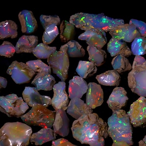 May include: A selection of raw opal gemstones, displaying a spectrum of colours such as blue, green, red, and yellow. The opals have a rough, natural texture and are set against a dark backdrop, accentuating their iridescent properties.