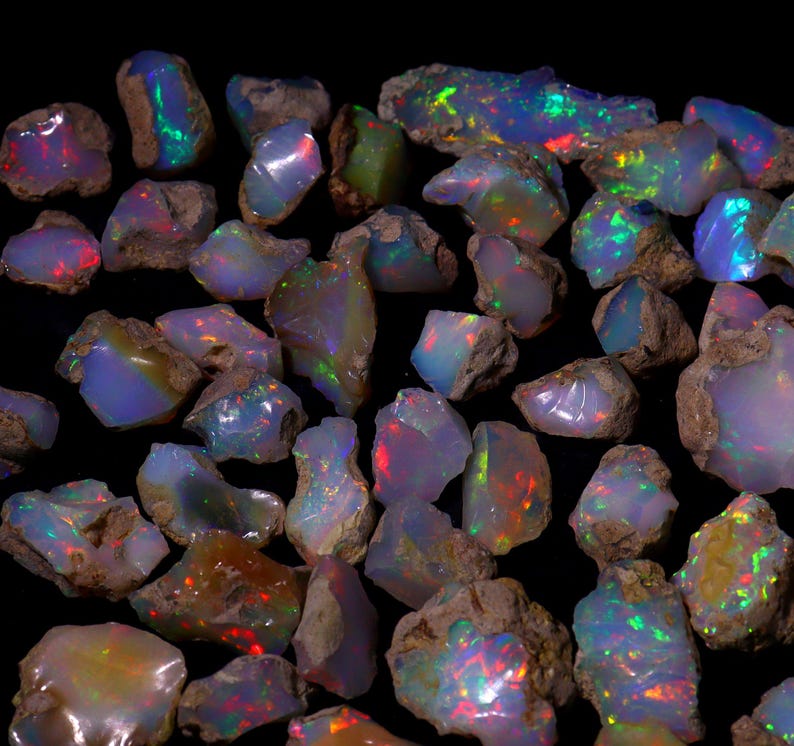 May include: A selection of raw opal gemstones, displaying a spectrum of colours such as blue, green, red, and yellow. The opals have a rough, natural texture and are set against a dark backdrop, accentuating their iridescent properties.
