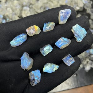 May include: A collection of raw, iridescent moonstone crystals. The stones display a range of colors, including blues, yellows, and whites. The crystals are rough-cut and vary in size, with a shimmering, natural appearance. The background shows more moonstones.