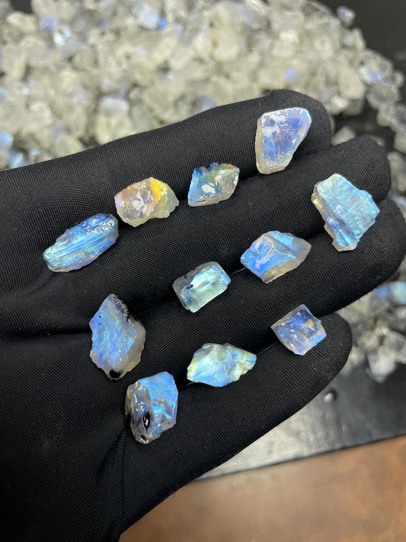 May include: A collection of raw, iridescent moonstone crystals. The stones display a range of colors, including blues, yellows, and whites. The crystals are rough-cut and vary in size, with a shimmering, natural appearance. The background shows more moonstones.