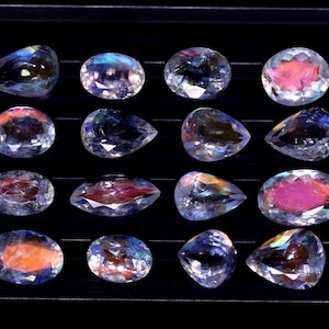 May include: Sixteen iridescent gemstones in various shapes, including oval and pear-cut. The gems display a range of colors, with pink, orange, and blue hues. The stones are arranged on a black surface, showcasing their facets and brilliance.