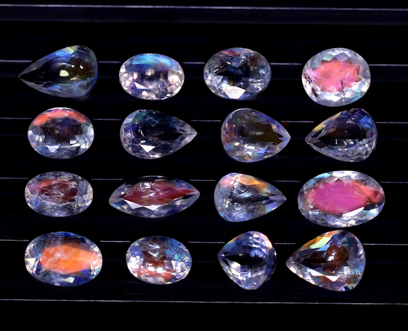 May include: Sixteen iridescent gemstones in various shapes, including oval and pear-cut. The gems display a range of colors, with pink, orange, and blue hues. The stones are arranged on a black surface, showcasing their facets and brilliance.