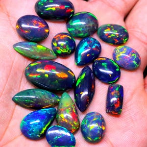 May include: A collection of 15 black opal gemstones with iridescent green, blue, and red flashes. The opals are various shapes and sizes, including oval, pear, and marquise.