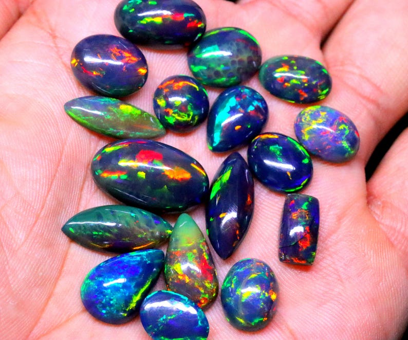May include: A collection of 15 black opal gemstones with iridescent green, blue, and red flashes. The opals are various shapes and sizes, including oval, pear, and marquise.