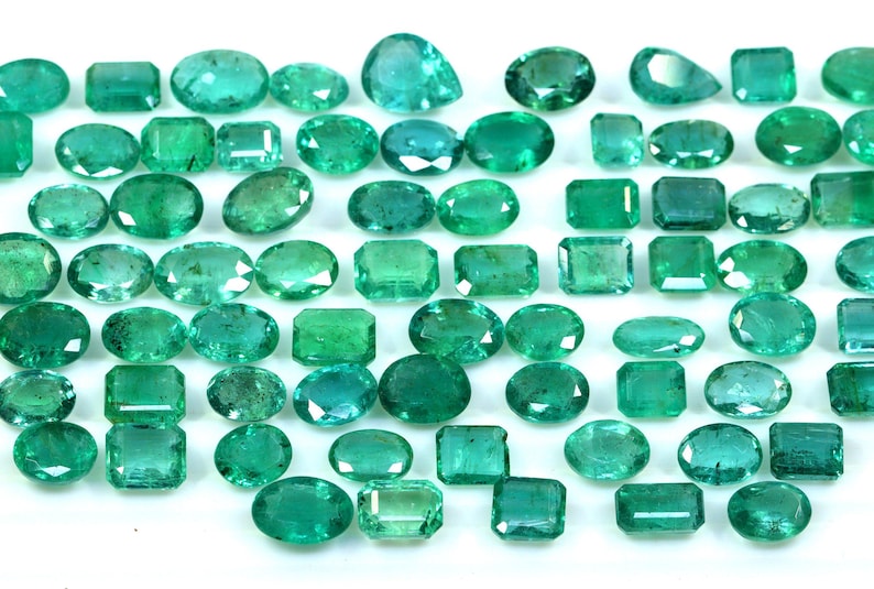May include: A collection of green emerald gemstones of various shapes and sizes, including oval, round, and rectangular.