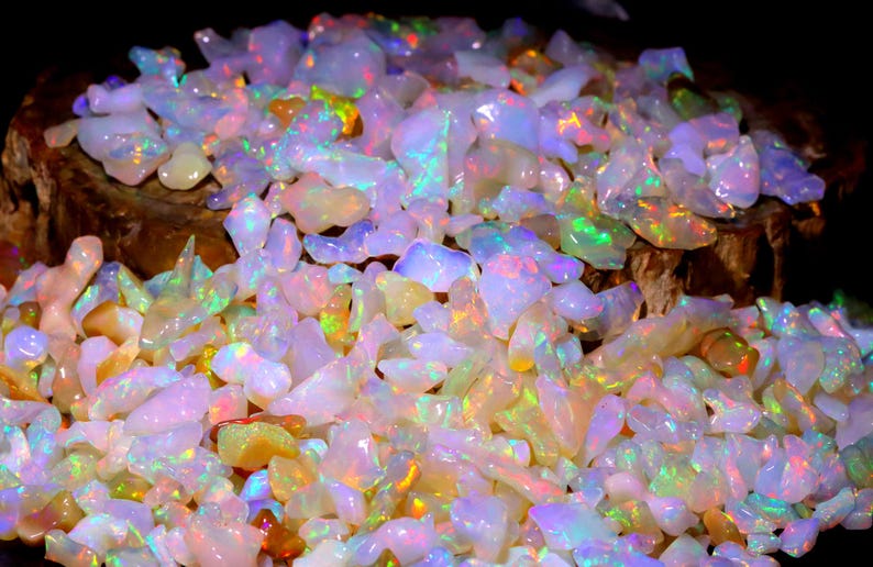 May include: A large pile of iridescent opal gemstones in various shapes and sizes. The opals are a mix of white, pink, and yellow with flashes of green and blue.