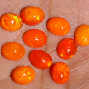 May include: A collection of ten oval-shaped, vibrant orange gemstones. The stones vary slightly in color and internal patterns, with some displaying a fiery, iridescent glow. The gemstones are displayed on a light-toned surface.