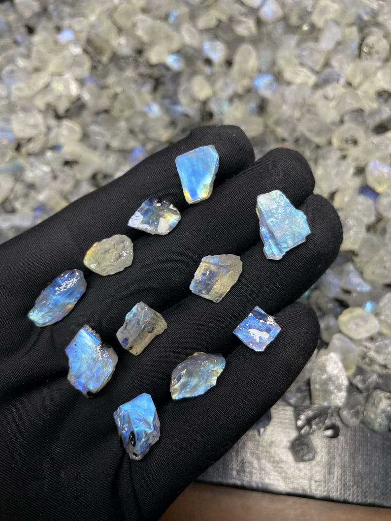 May include: A collection of raw, iridescent moonstone gemstones. The stones, in various shapes and sizes, display a range of colors including blues, yellows, and whites. The gemstones are held in a black gloved hand, with a blurred background of similar stones.
