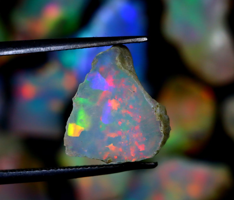May include: A rough, iridescent opal gemstone with a white base and flashes of green, blue, red, and yellow. The opal is held by tweezers.