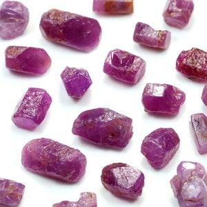 May include: A collection of pink ruby crystals, each with a unique shape and size. The crystals are arranged on a white surface.