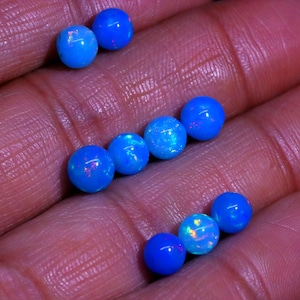 May include: A collection of eight blue opal beads, each with a unique iridescent shimmer. The beads are round and smooth, and they are arranged in a loose pile on a hand.