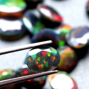 Op de afbeelding: Een close-up van een donkergroene, zwarte en rode ovale opaal edelsteen die met een pincet wordt vastgehouden. De edelsteen wordt omringd door andere opalen in vergelijkbare kleuren.