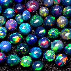 May include: A collection of round, iridescent opal gemstones in various shades of blue, green, and red. The opals are arranged in a random pattern on a black surface.