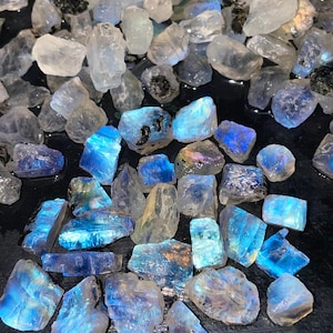 May include: A collection of raw, uncut moonstone crystals. The stones vary in size and shape, with a mix of clear, white, and blue hues. Some stones have a striking blue flash. The crystals are scattered on a dark surface.