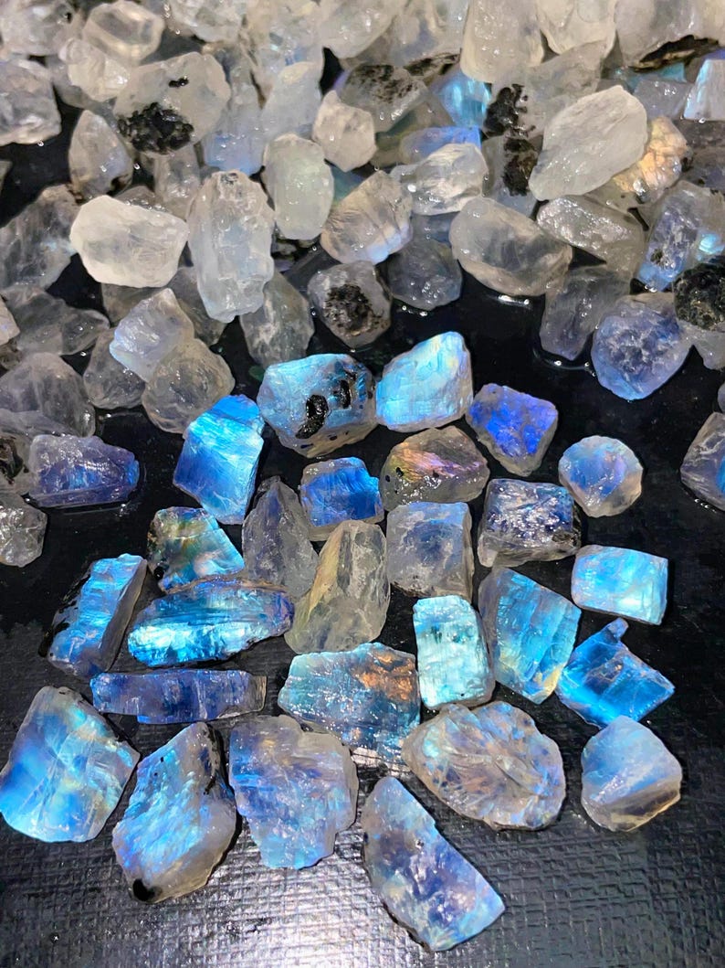 May include: A collection of raw, uncut moonstone crystals. The stones vary in size and shape, with a mix of clear, white, and blue hues. Some stones have a striking blue flash. The crystals are scattered on a dark surface.