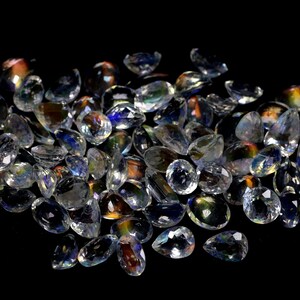 May include: A collection of clear, faceted gemstones with iridescent flashes of color. The gemstones are various shapes, including oval and teardrop, and are scattered on a black surface. The light reflects off the facets, creating a sparkling effect.