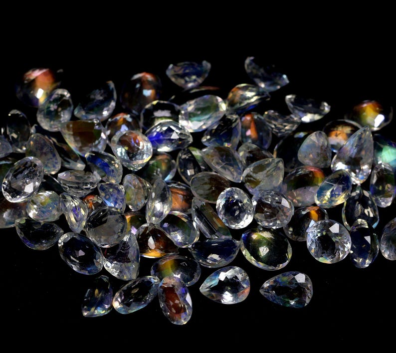 May include: A collection of clear, faceted gemstones with iridescent flashes of color. The gemstones are various shapes, including oval and teardrop, and are scattered on a black surface. The light reflects off the facets, creating a sparkling effect.