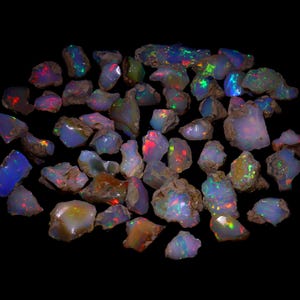 May include: A selection of raw opal gemstones, showcasing a spectrum of colours such as blue, green, red, and yellow. The opals are in a rough-cut state, with a natural, unpolished look. The gemstones are arranged against a black backdrop.