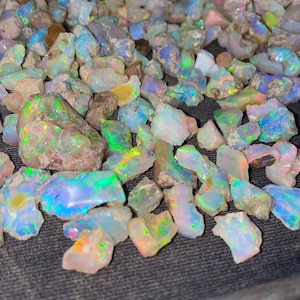 Op de afbeelding: Een verzameling ruwe, iriserende opaal edelstenen in verschillende maten en vormen. De opalen zijn een mix van witte, blauwe, groene en roze tinten.