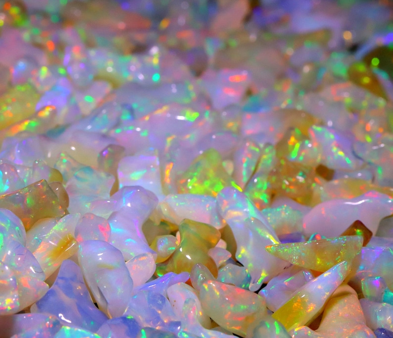 May include: A close-up view of a pile of iridescent opal gemstones. The opals are various shades of white, pink, and green, with a shimmering, rainbow-like effect.