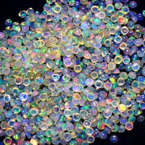 Op de afbeelding: Een grote stapel iriserende opaal cabochons in verschillende maten. De opalen zijn een mix van witte, heldere en regenboogkleuren.