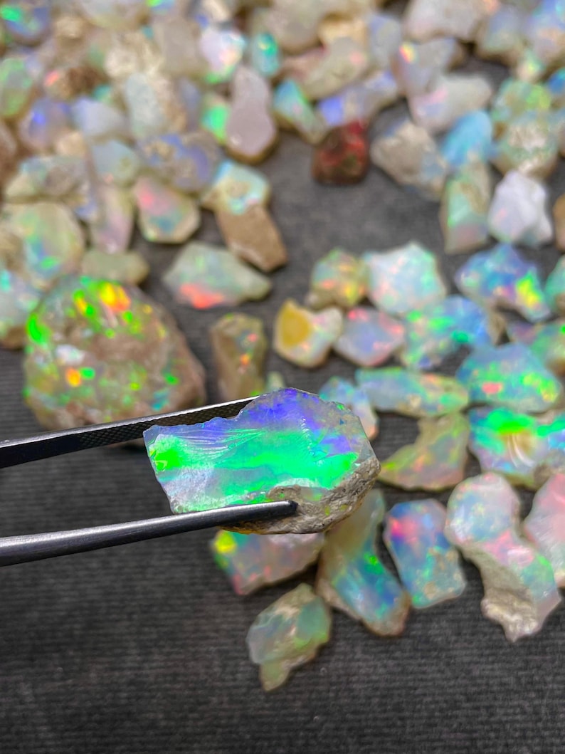 Op de afbeelding: Een verzameling ruwe opaal edelstenen, met een grote opaal die met een pincet wordt vastgehouden. De opaal is iriserend met groene, blauwe en witte kleuren.