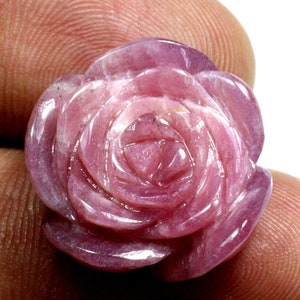 May include: A carved rose made of pink gemstone. The rose has detailed petals and a polished surface. The gemstone is a light pink color with darker pink veining. The rose is held in a person's hand.