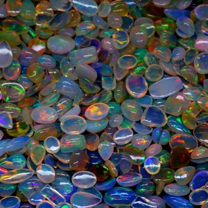 Op de afbeelding: Een verzameling iriserende opaal edelstenen in verschillende vormen en maten. De opalen vertonen een scala aan kleuren, waaronder blauw, groen, geel en rood, met een glinsterende, doorschijnende kwaliteit. De edelstenen zijn verspreid.