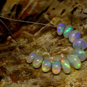 May include: A strand of oval-shaped opals, each displaying iridescent colors of green, blue, and orange. The opals are strung together on a thin, dark string, set against a textured, brown background.