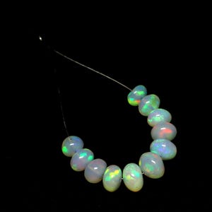 May include: A strand of oval-shaped opal beads. The opals display a milky white base color with flashes of green, yellow, and red. The beads are strung on a thin, nearly invisible thread against a black background.