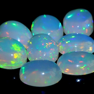 Könnte beinhalten: Acht ovale Opale mit milchig-weißer Grundfarbe und schillernden grünen, blauen und roten Reflexen. Die Edelsteine sind vor einem schwarzen Hintergrund angeordnet, wodurch ihr lebendiges Farbspiel hervorgehoben wird.